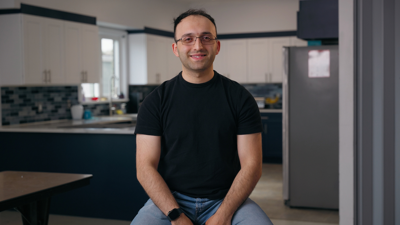 Man smiling with glasses and a black shirt and jeans sitting in a kitchen