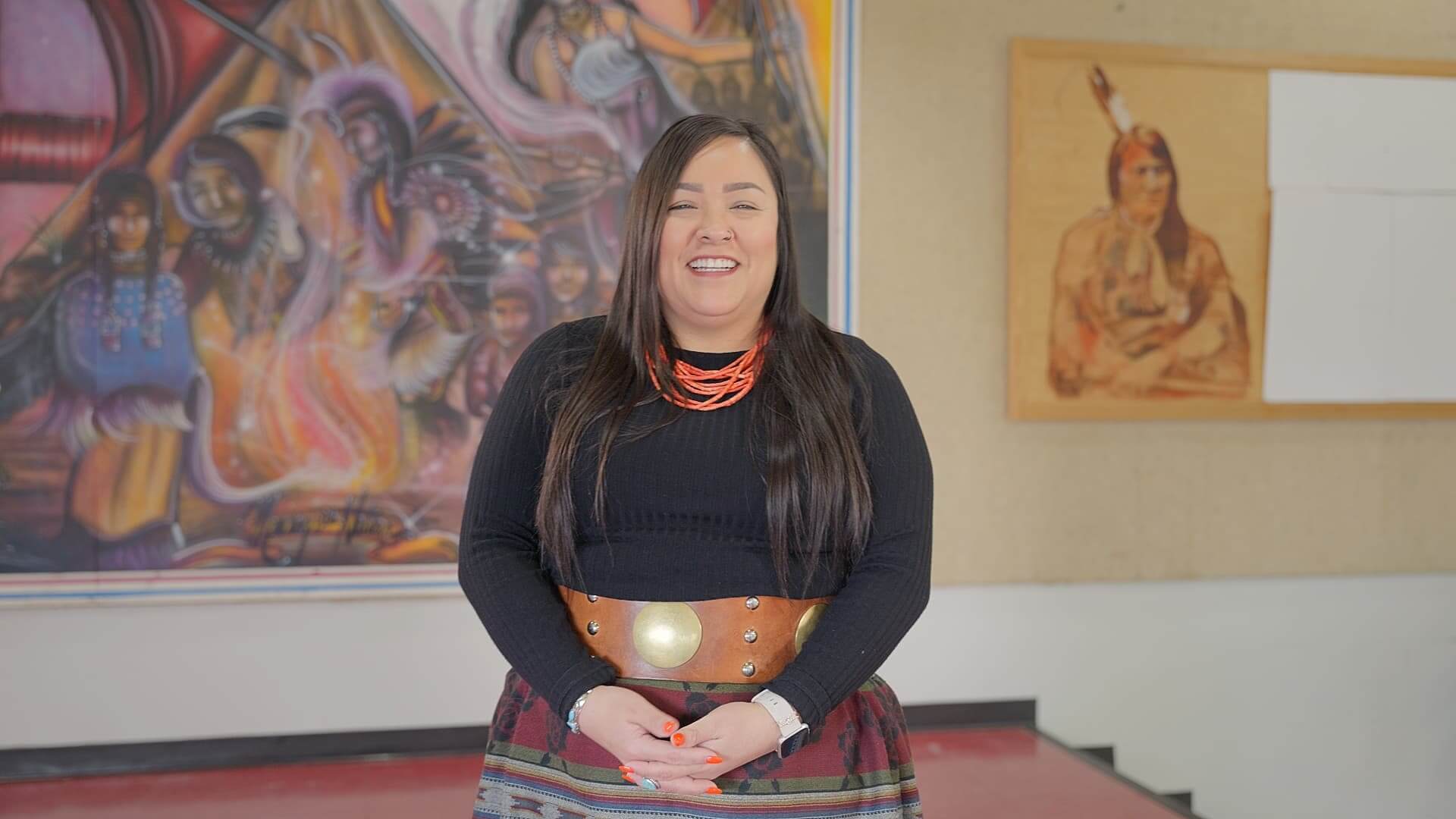 Woman smiling in black top and wide brown belt standing in front of a colorful mural