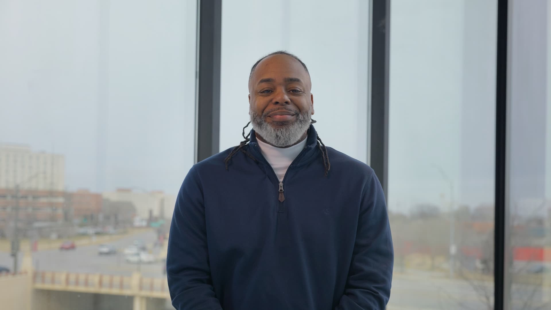 A man with dreadlocks and a white beard wearing a navy blue halfzip sweater standing in front of a large window