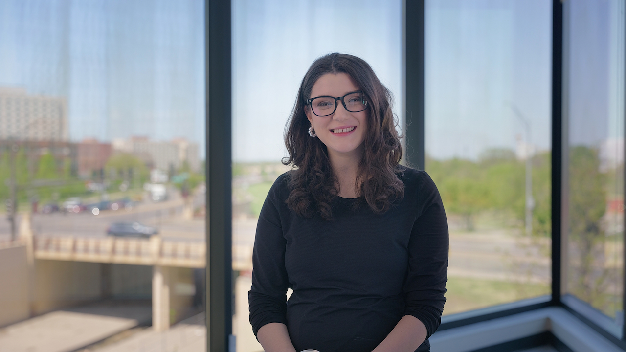 Smiling woman with dark hair and glasses sitting in front of a large window