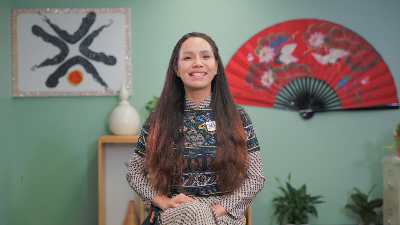Smiling woman with long dark hair in a colorful patterned shirt sitting in front of a wall with a large red accordion collapsible fan and a self with figurines behind her